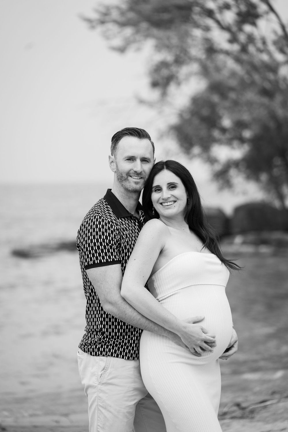 Black and white romantic beach photo series shows a couple embracing and smiling together in casual attire.