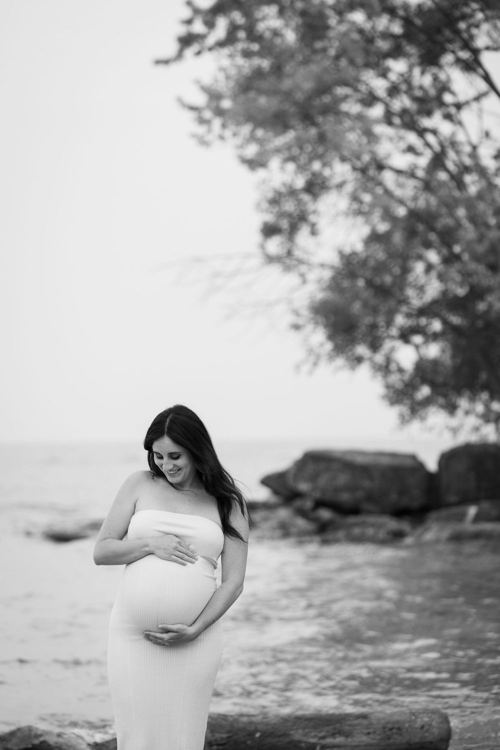 A black and white photo of someone in a flowing white gown cradling their pregnant belly by a rocky shore.