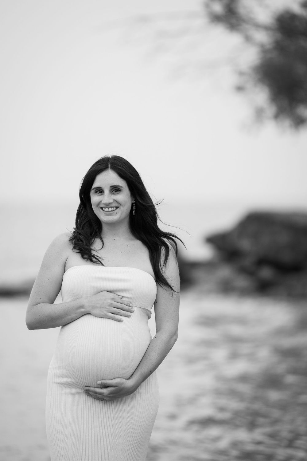 Black and white maternity photography series shows an expectant mother in a white dress against a soft blurred background.