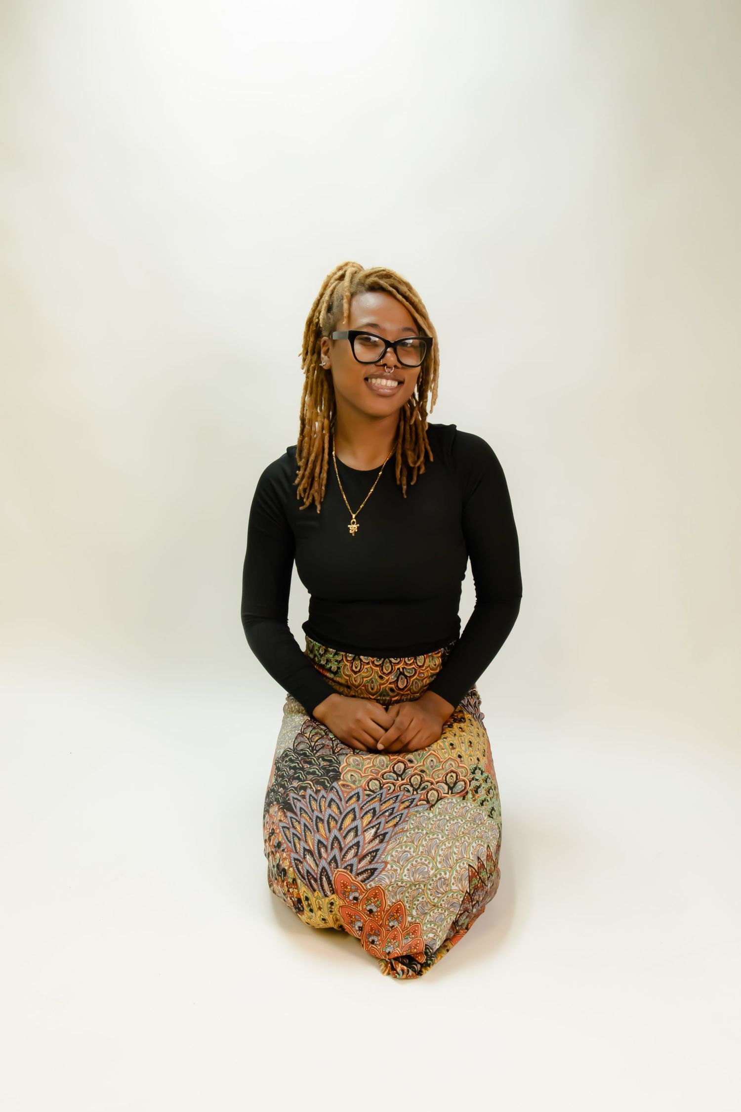 Seated figure wearing black long sleeve top and patterned skirt against white background in studio portrait setting.