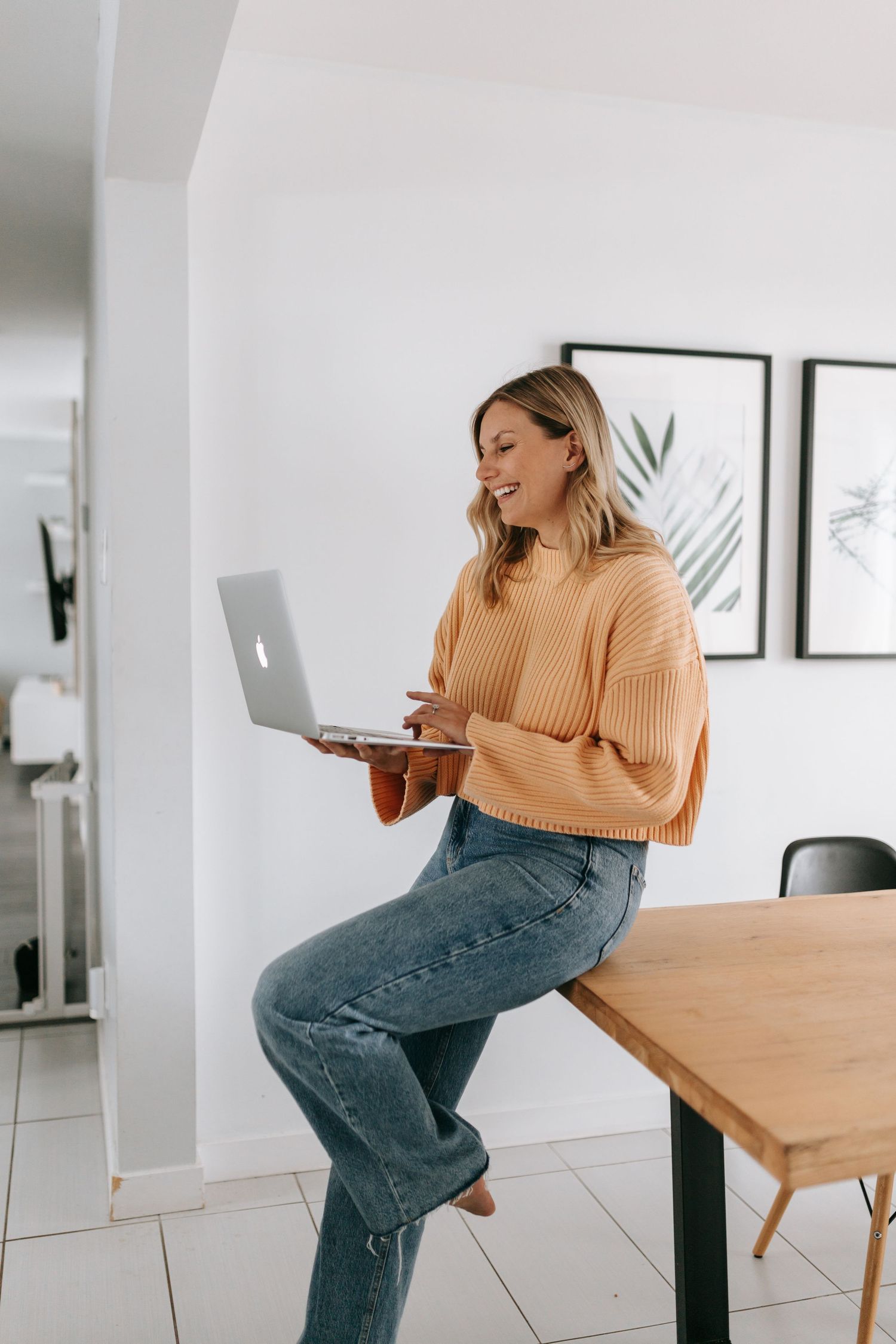Happy woman in orange sweater using laptop while sitting on wooden table at home.