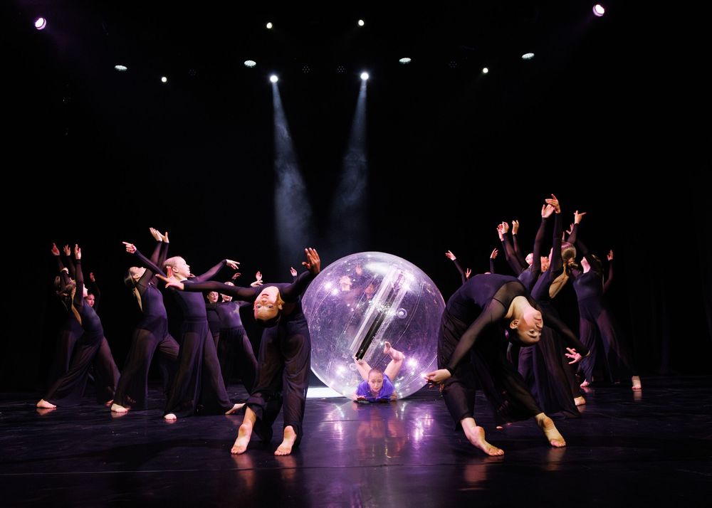 Performers in black costumes dance around a large transparent sphere on a dark stage with dramatic lighting.