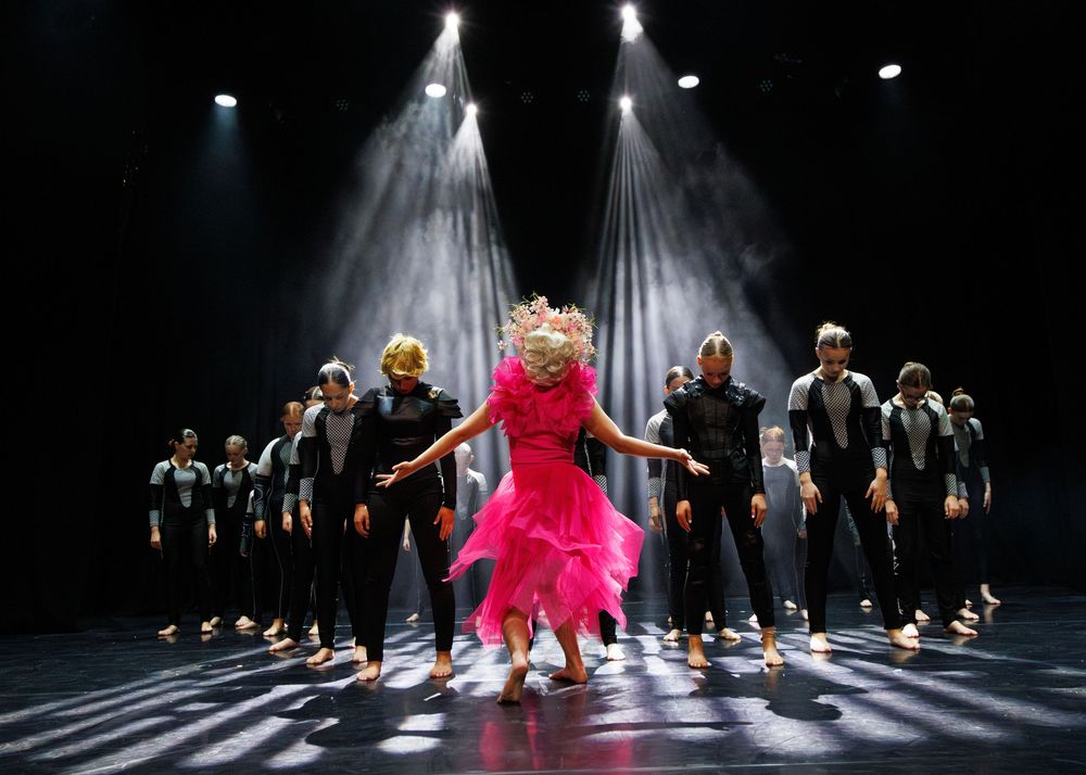 Dancers in black perform on a dark stage behind a figure in a bright pink dress under dramatic spotlights.