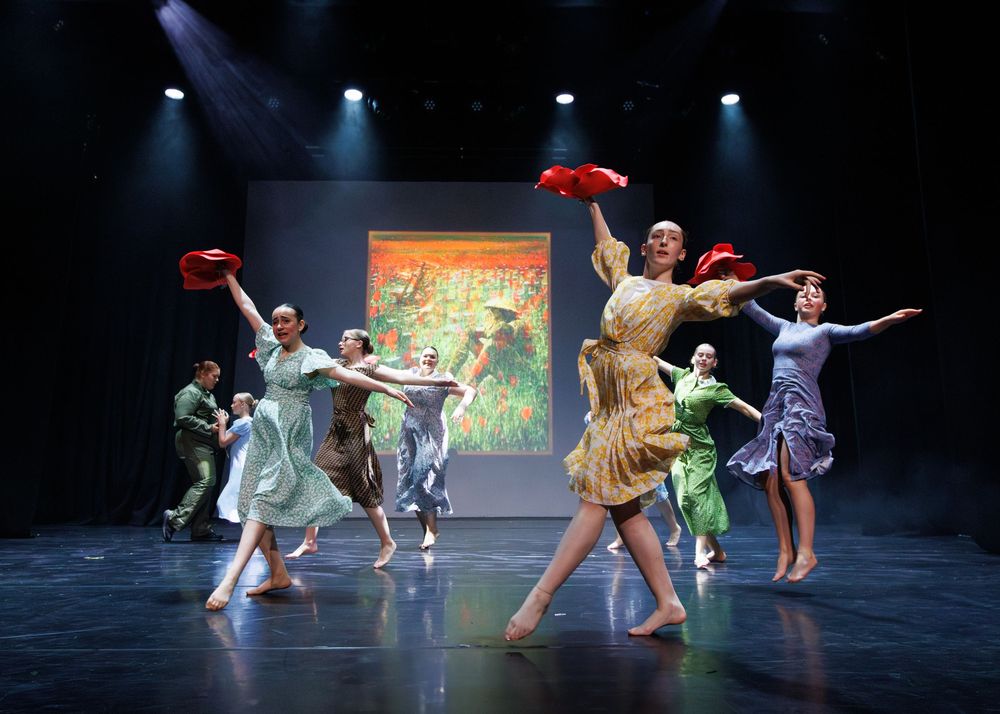 Dancers in colorful costumes perform on a dimly lit stage with artistic lighting and backdrop during a theatrical show.