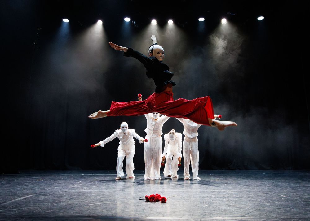 Dramatic dance performance on stage with a figure in red leaping above white-clad dancers amid theatrical lighting.