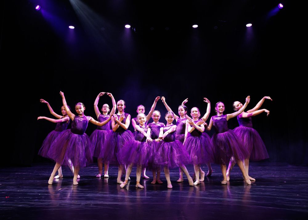 Ballet dancers in purple tutus perform synchronized movements on a dark stage illuminated by blue spotlights.