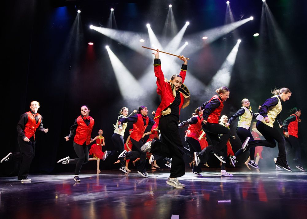 Dance performers in red jackets execute a synchronized lean move during a dynamic stage performance under dramatic lighting.