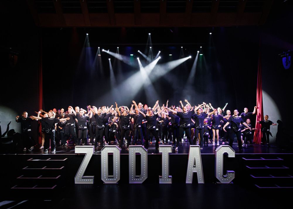 Orchestra performers on stage with dramatic spotlights and large ZODIAC text displayed during a theatrical production.