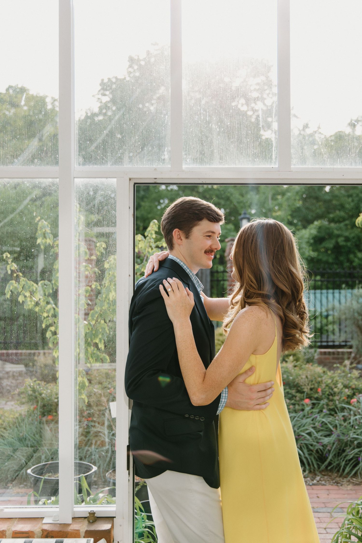 A couple shares a joyful dance together in a bright sunlit room with large windows and lush garden views outside.