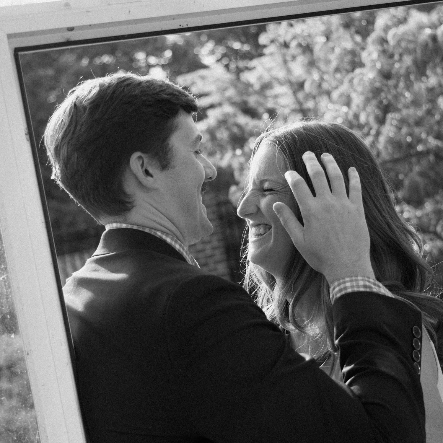 A romantic black and white portrait of a couple sharing an intimate moment by a window during an engagement photoshoot.