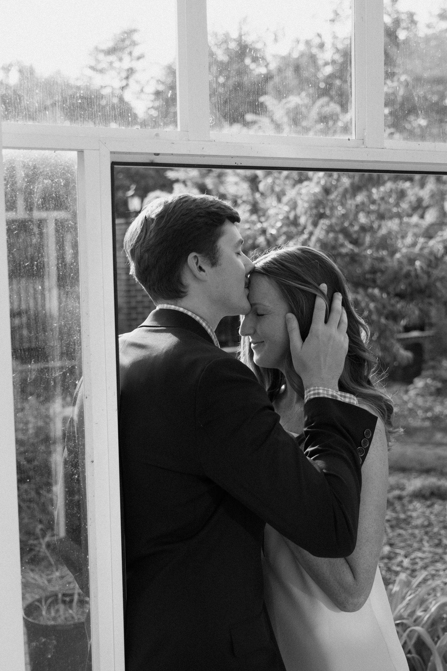 A romantic black and white photograph of a tender embrace near a window with garden scenery in the background.