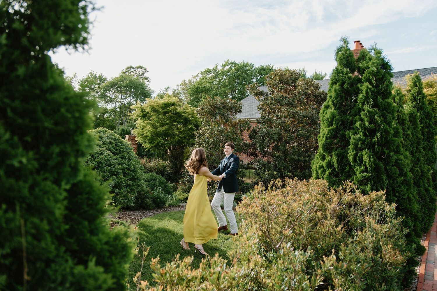 A couple shares a romantic dance moment together in a lush garden setting surrounded by evergreen trees and bushes.