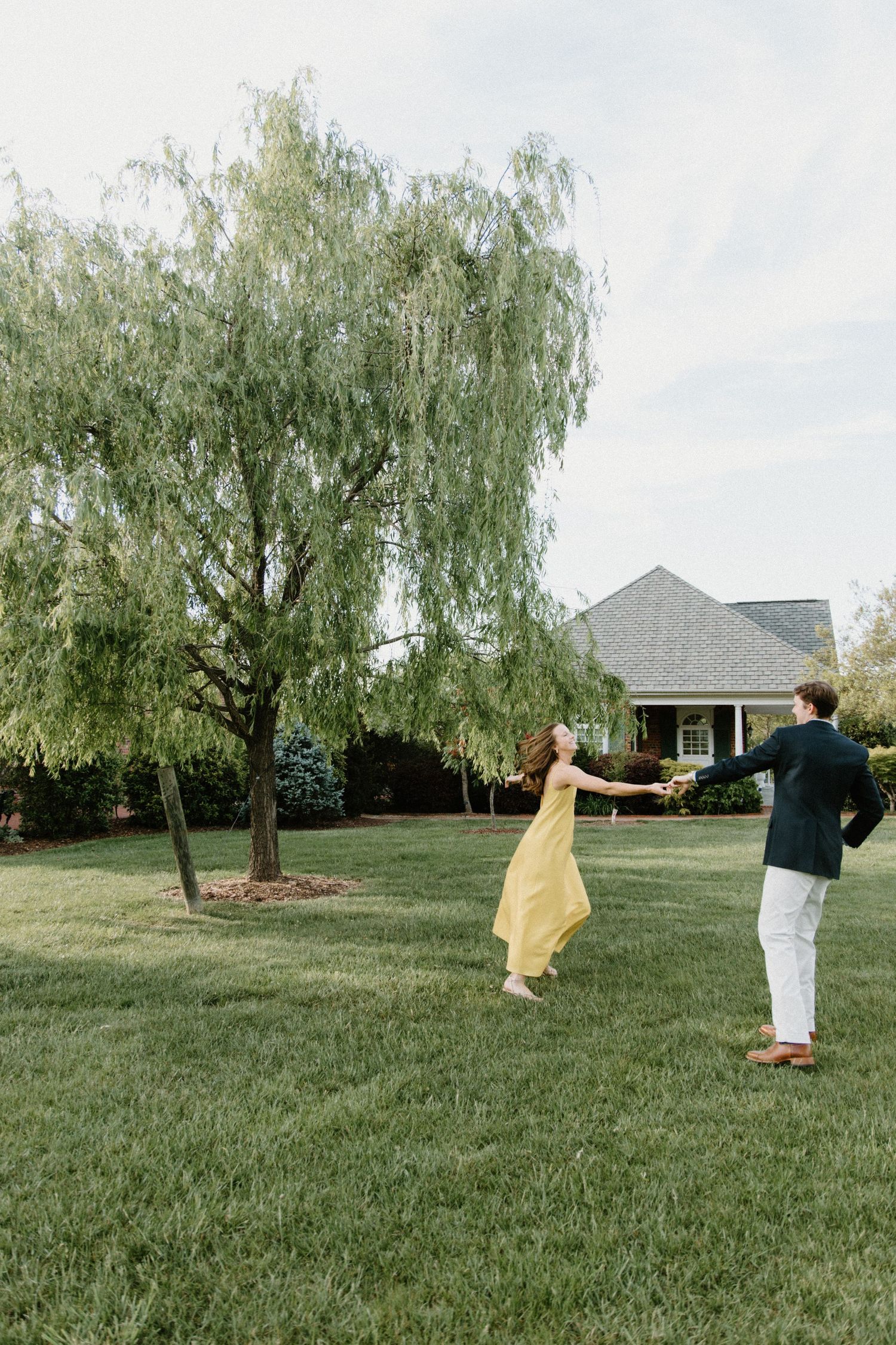 Two people dance playfully on a green lawn with a weeping willow tree and house in the background.