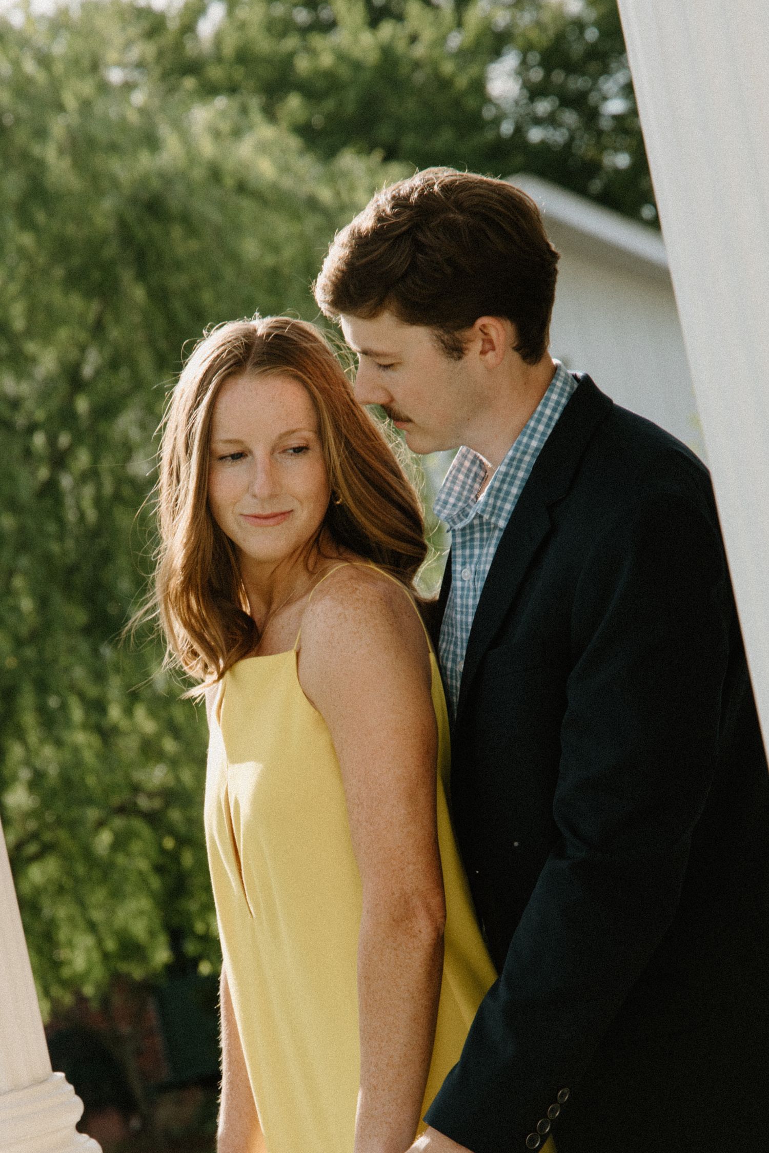 A couple in formal wear stand close together against a sunlit outdoor backdrop with white pillars.