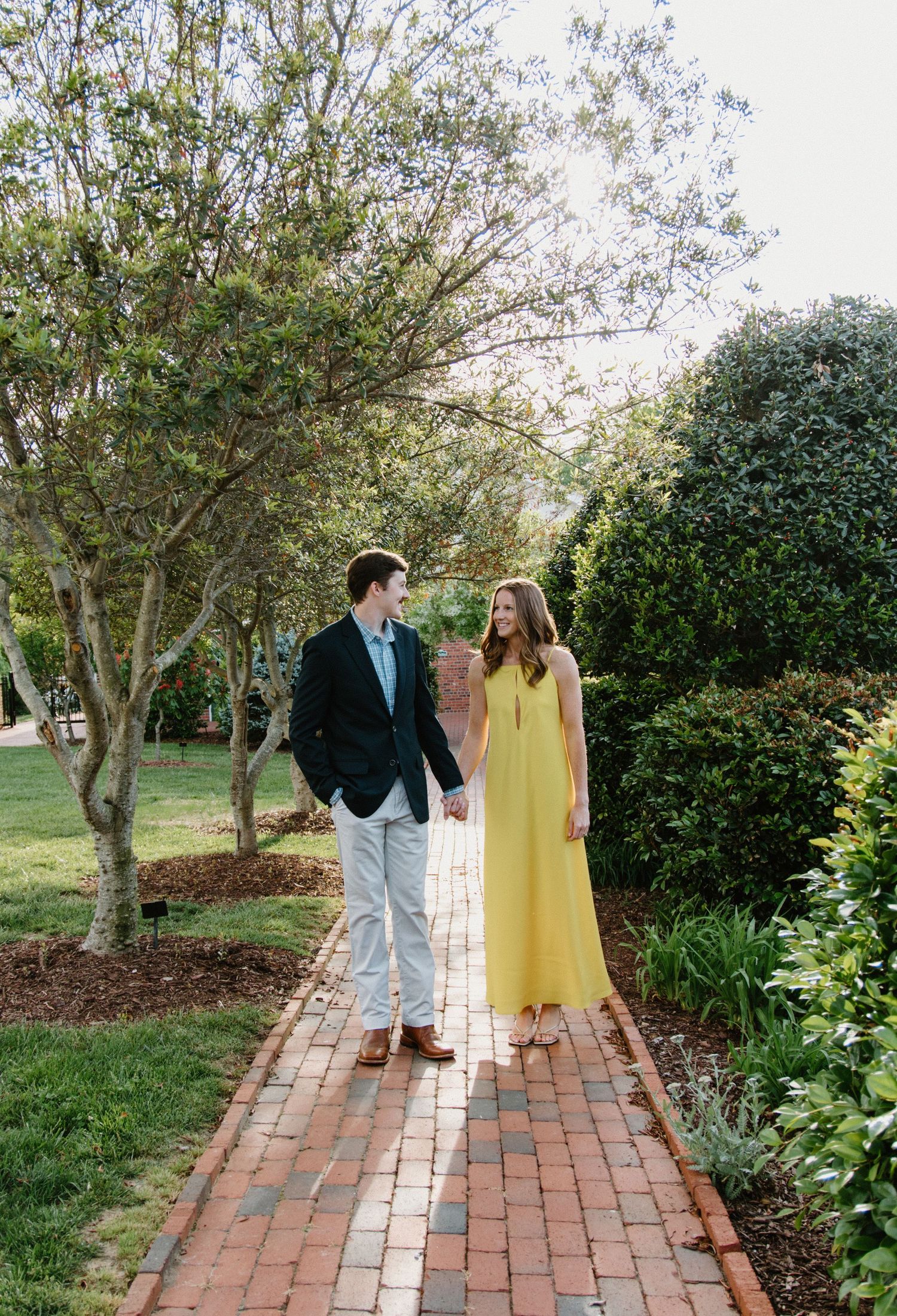 A couple strolls along a scenic brick pathway surrounded by lush gardens and trees at an outdoor venue.