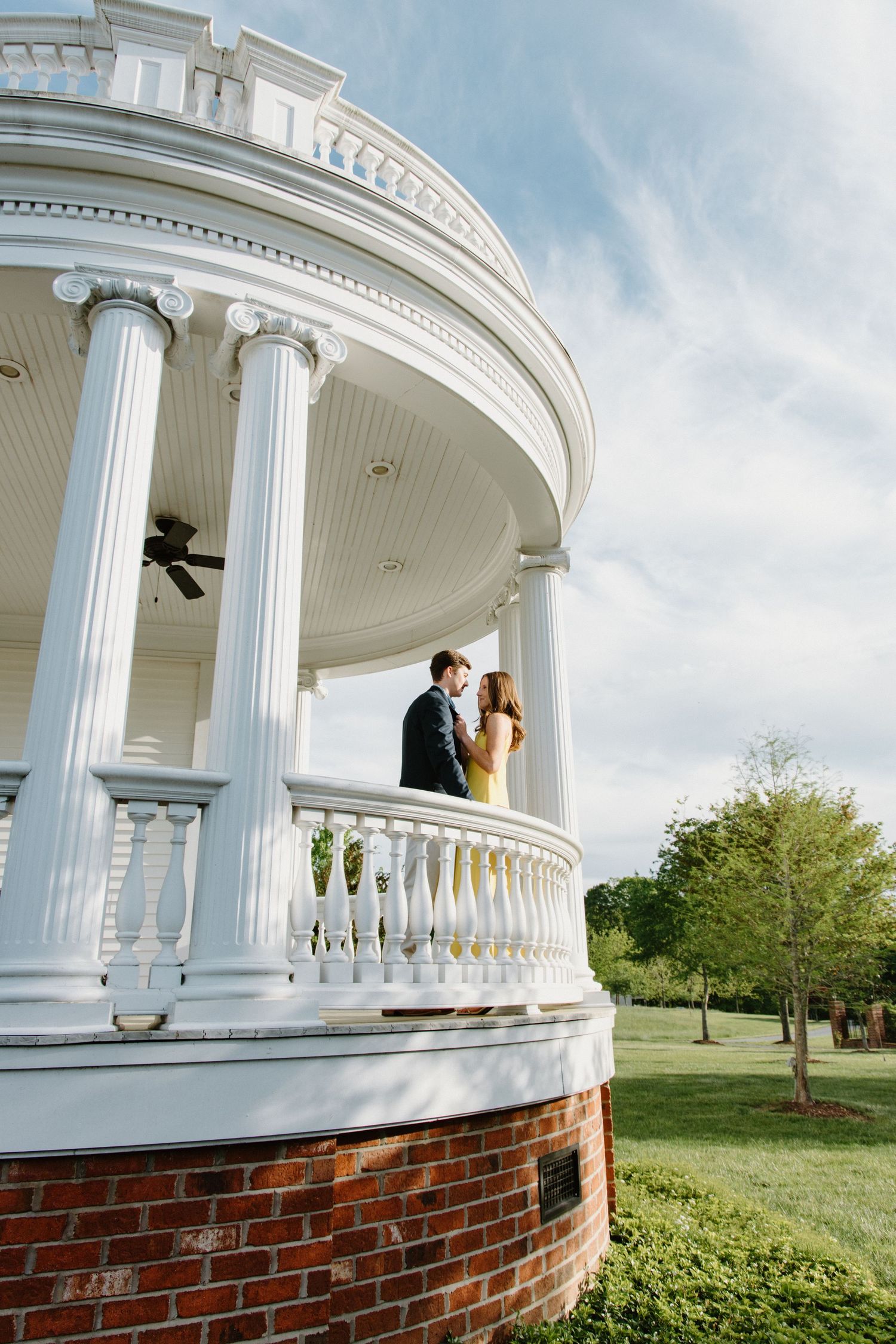 A white columned gazebo with brick foundation sits elegantly on a lawn against a blue sky.