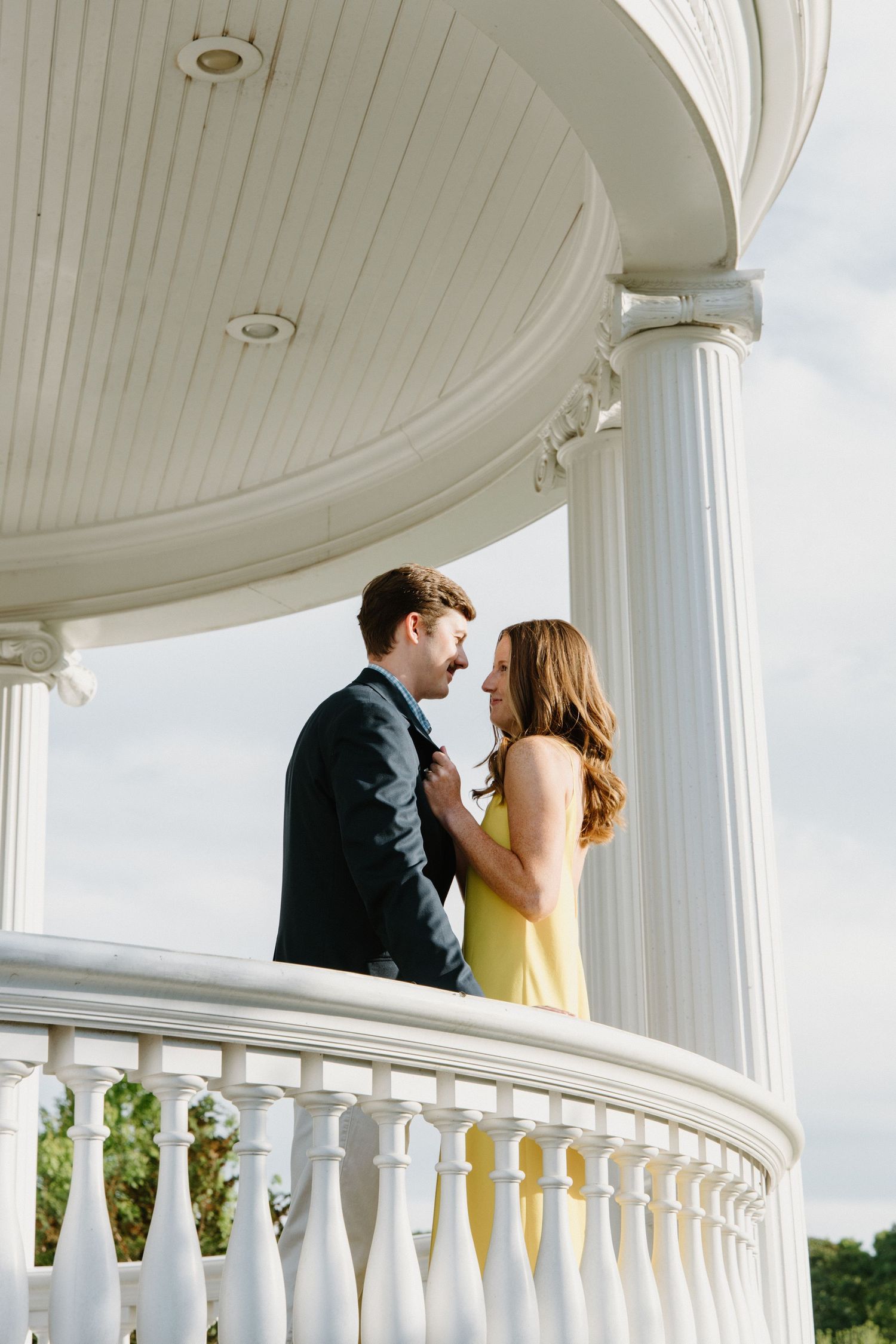 Couple sharing a romantic moment on an elegant white columned balcony with a yellow dress catching sunlight.