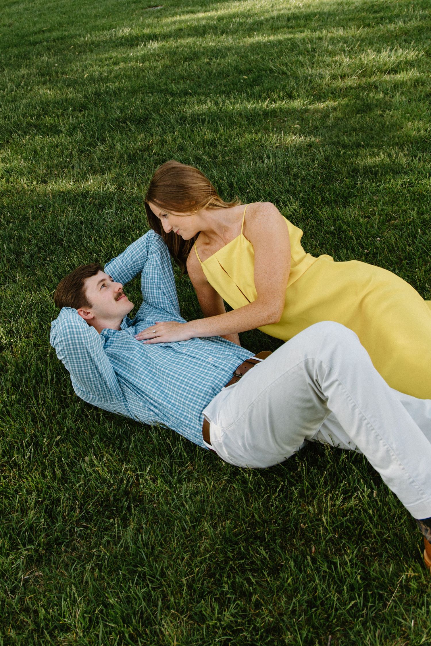 Two people in casual attire embrace while lying on green grass on a sunny day.