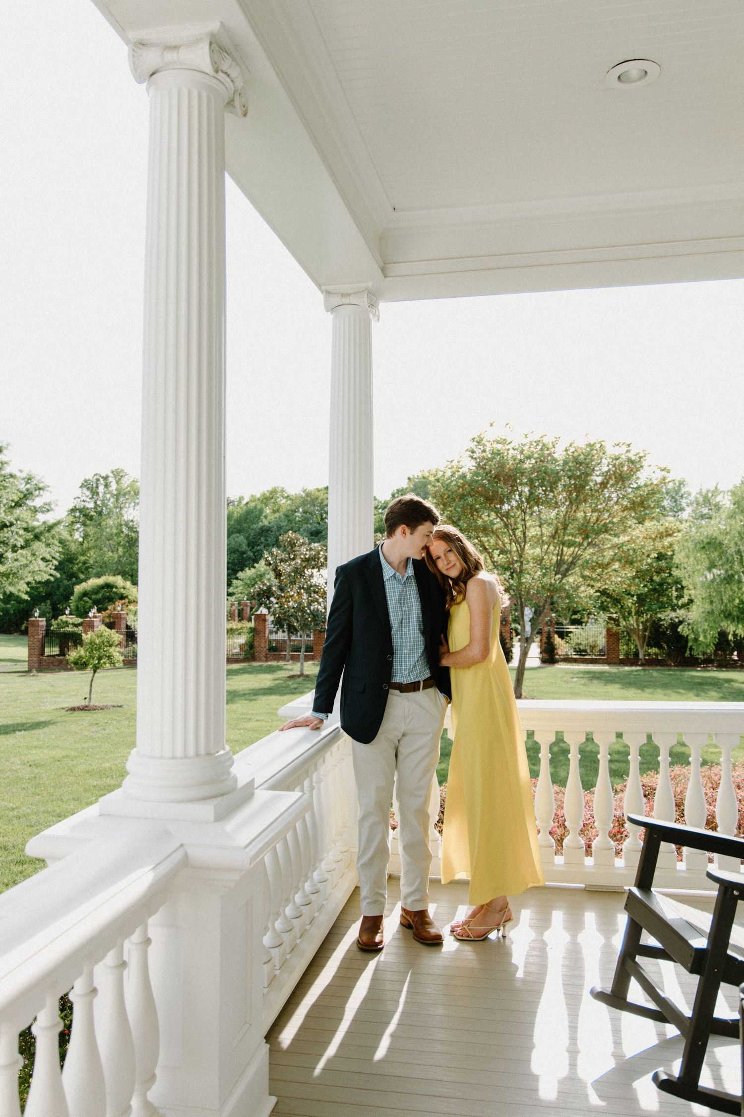 A couple shares an intimate moment on an elegant white columned porch with lush greenery in the background.