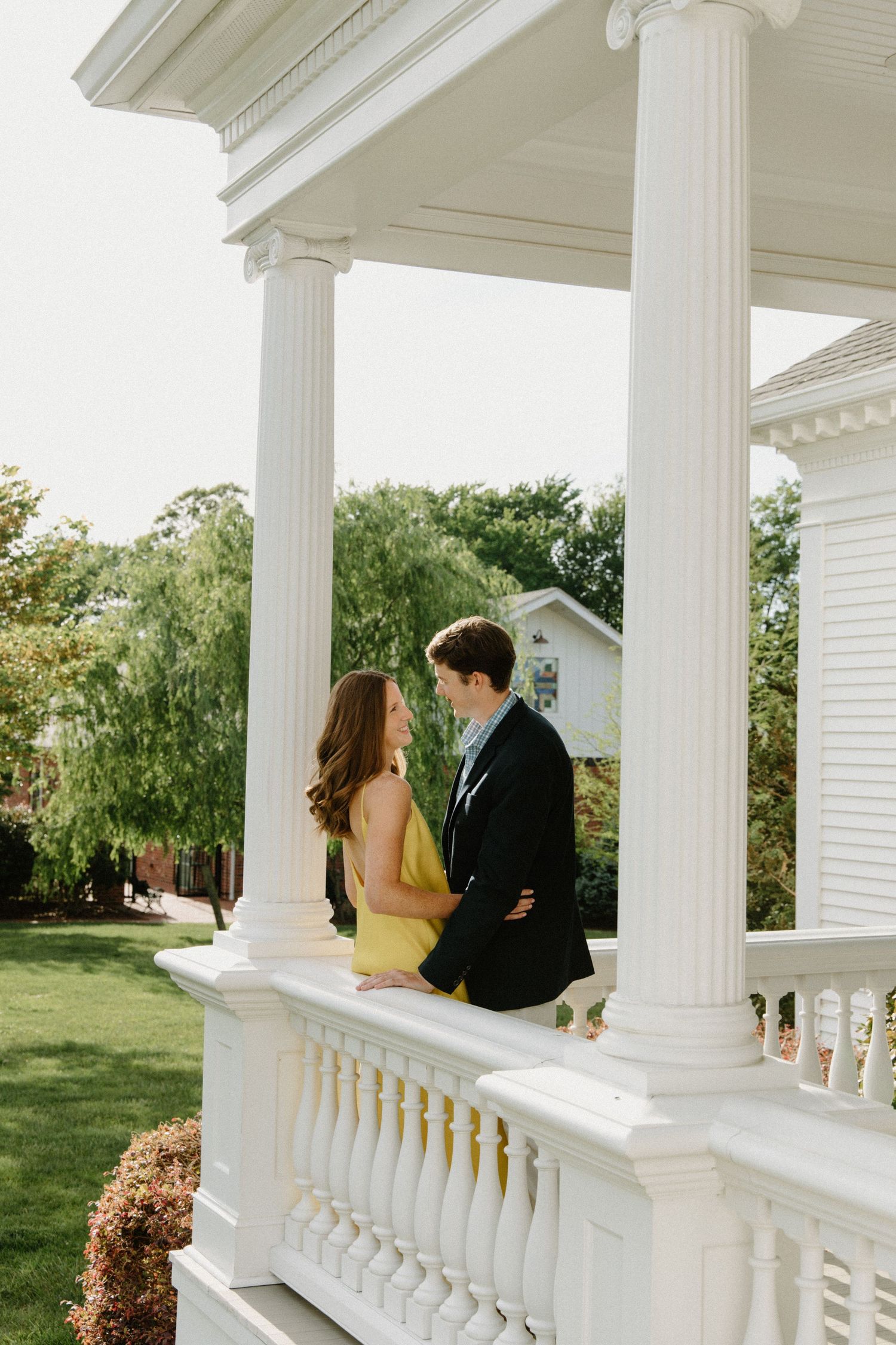 A couple shares a tender moment on an elegant white-columned porch with greenery in the background.
