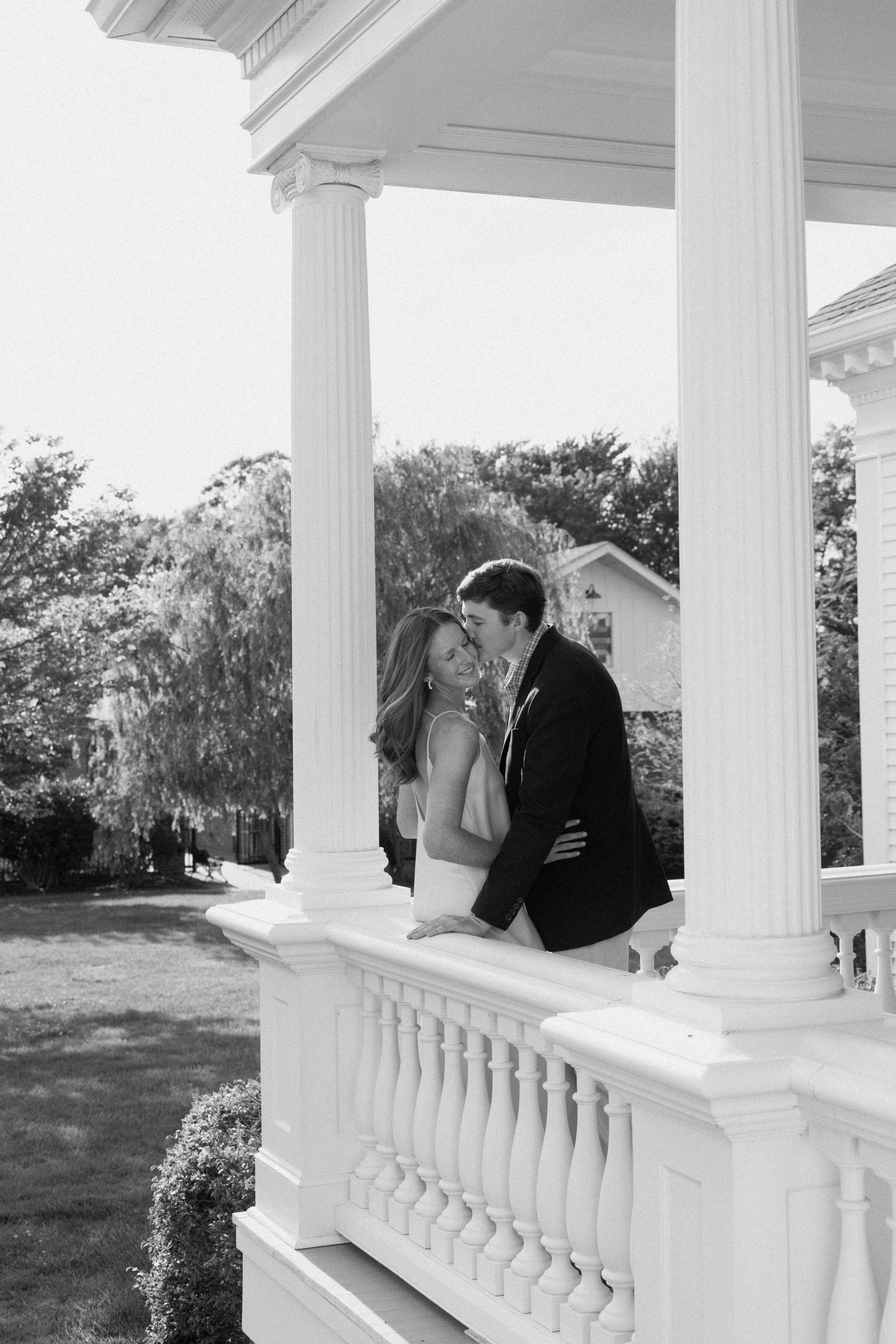 A romantic black and white photo of a couple embracing on an elegant white columned porch of a southern-style mansion.