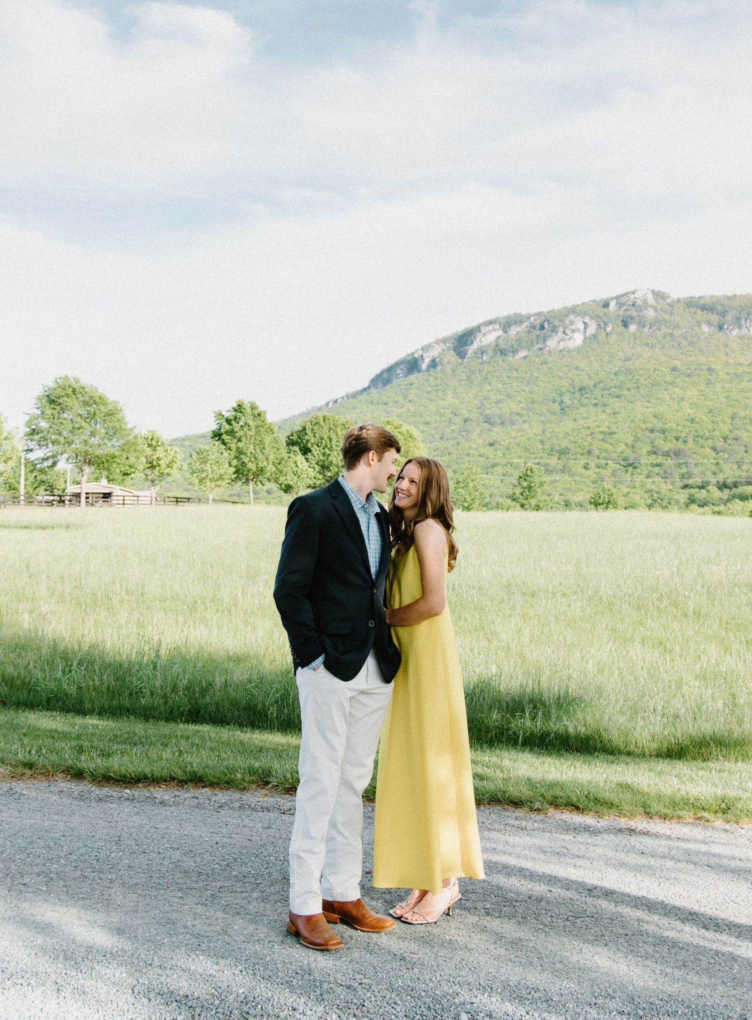 A couple embraces on a country road with a scenic mountain landscape and green meadow in the background.