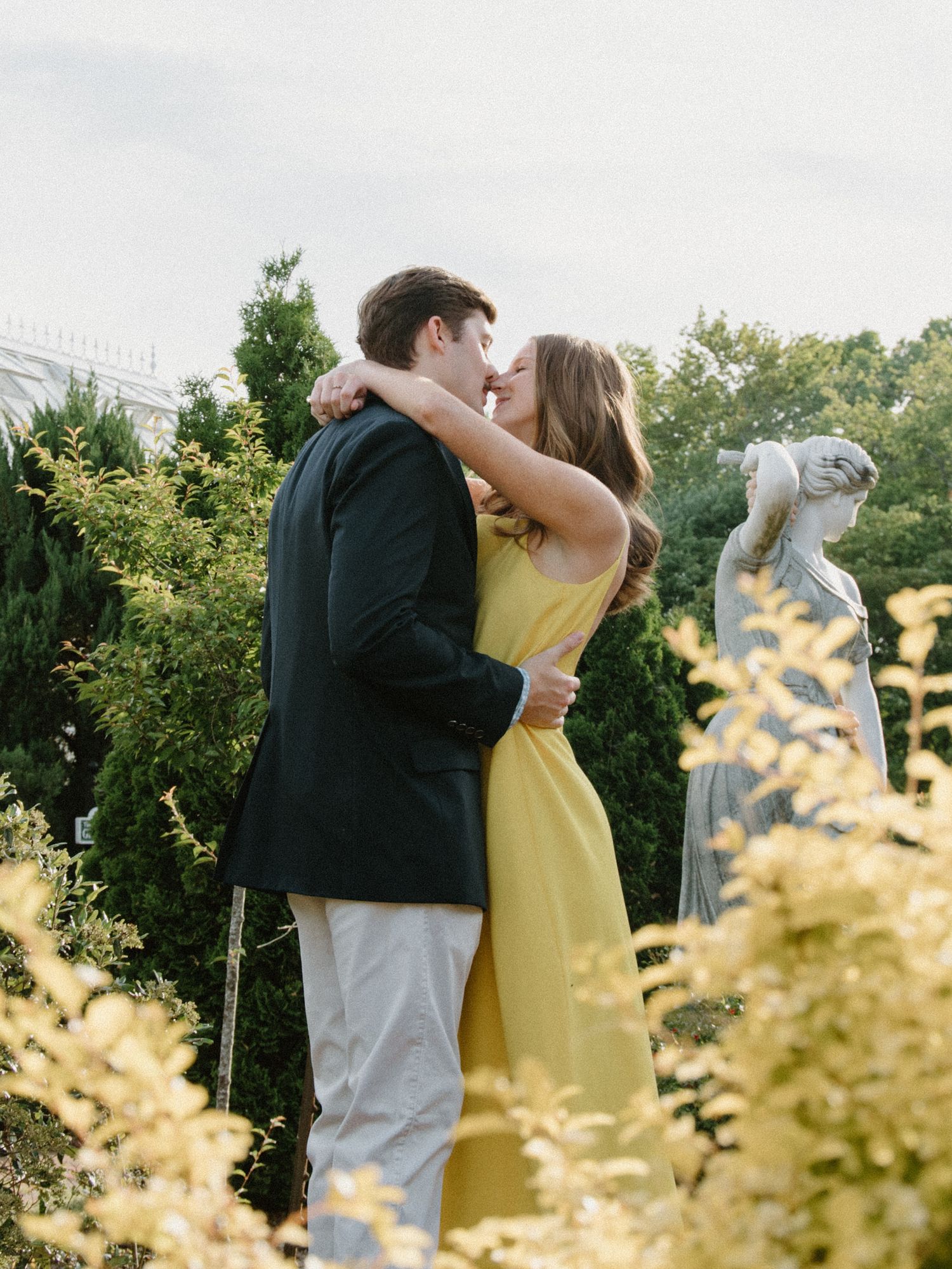 A romantic couple embracing in a garden with yellow flowers and a statue in the background at dusk.