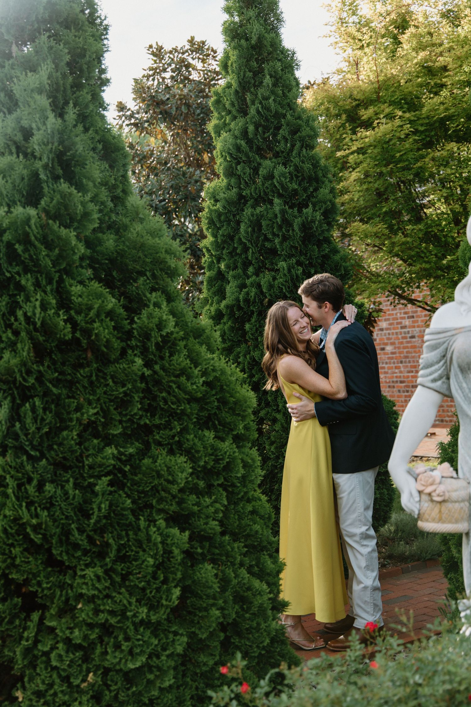 A couple shares an intimate moment among tall evergreen trees and brick walls in a garden setting.