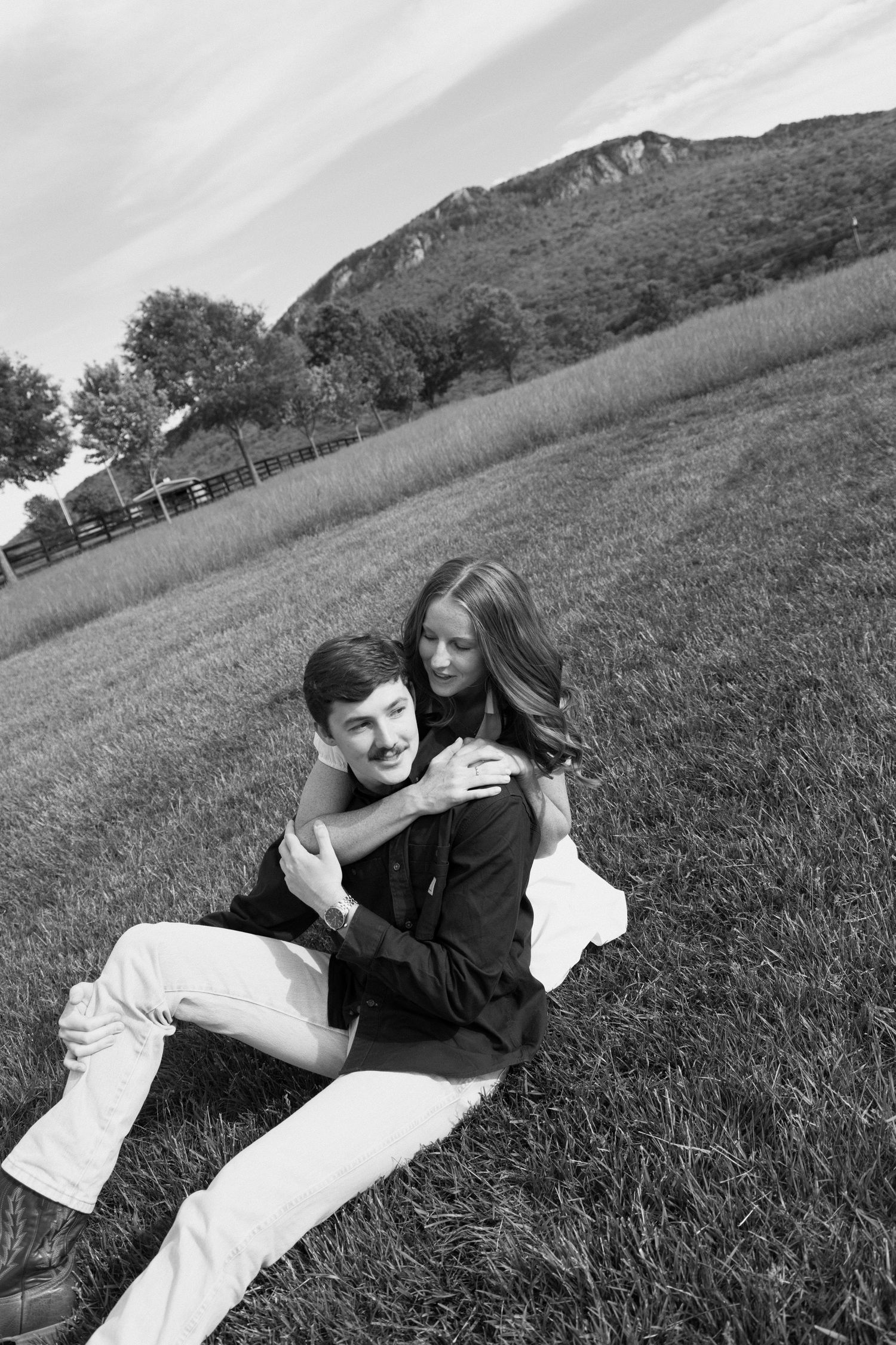 A couple laughs together while laying on a grassy hillside with mountains in the background in black and white.