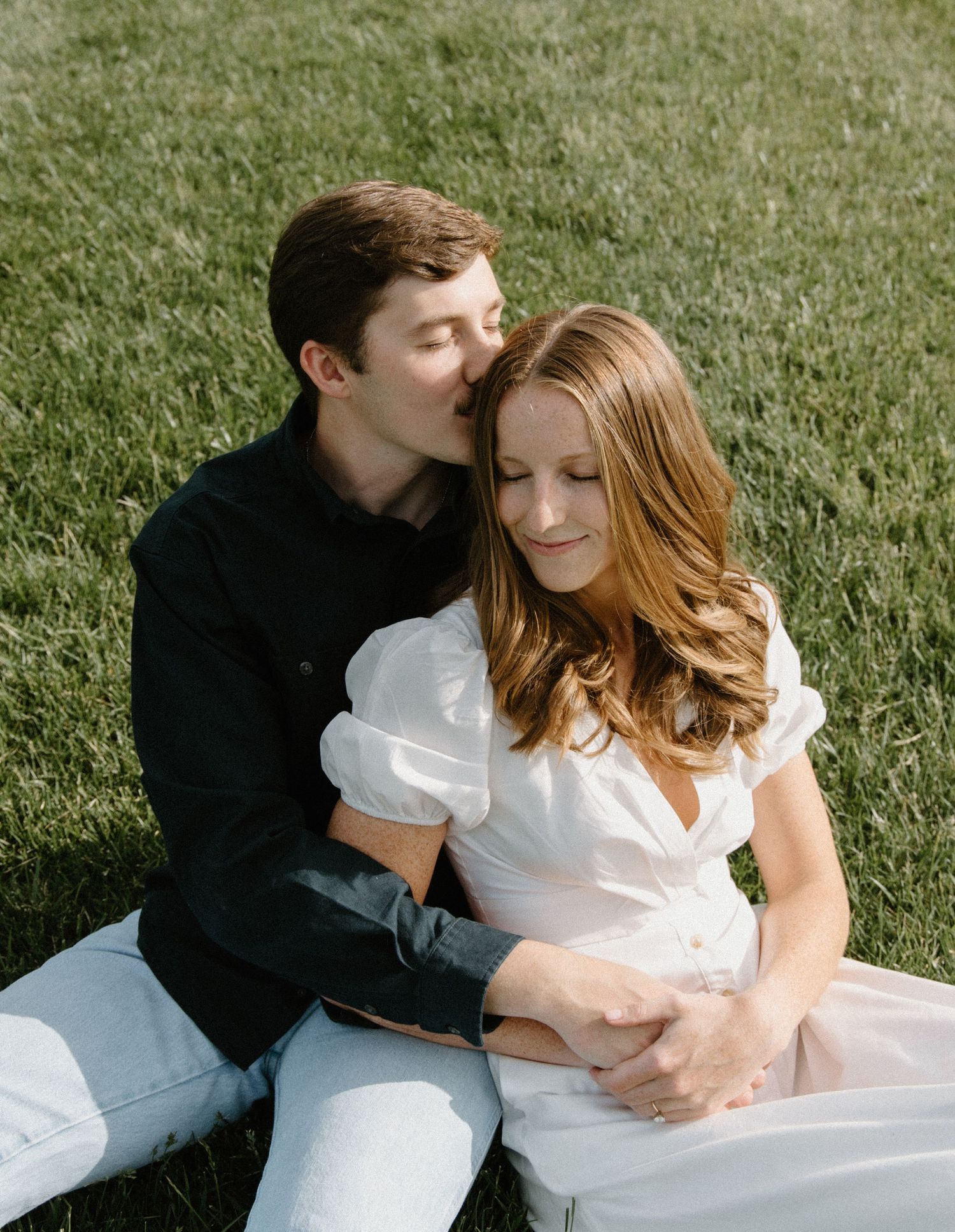 Couple sits intimately on grass with woman in white dress and man in black shirt in romantic outdoor setting.