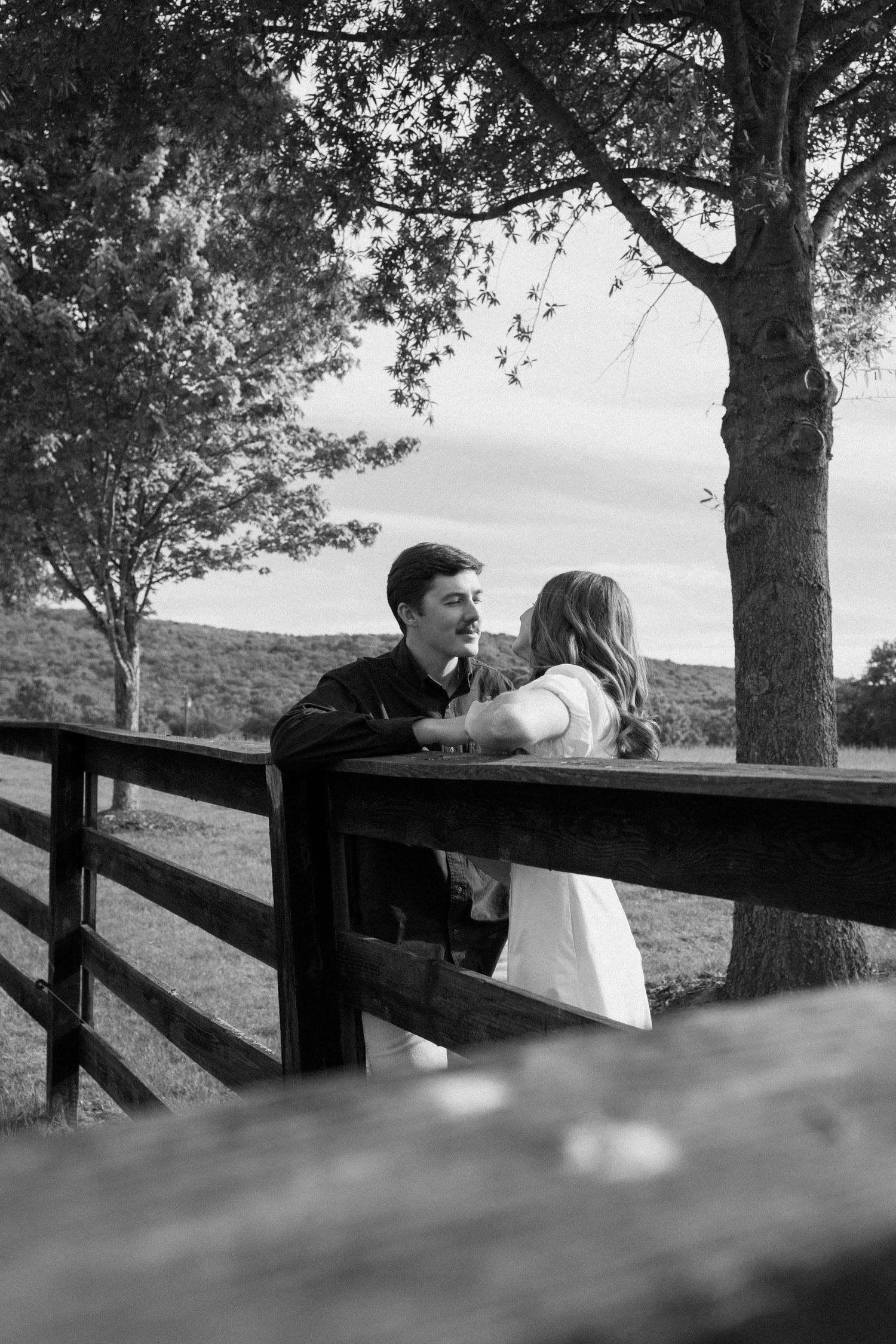 A couple shares an intimate moment while leaning against a wooden fence in a scenic black and white landscape.