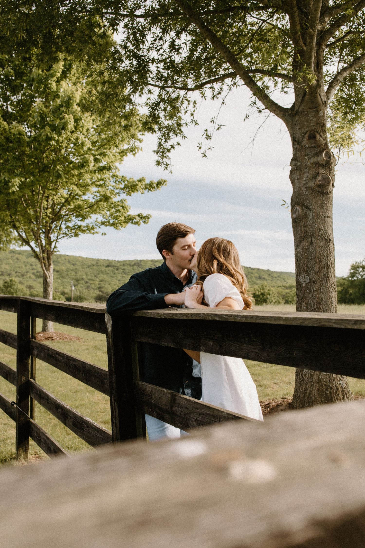 A couple shares an intimate moment leaning on a wooden fence overlooking rolling hills on a sunny afternoon.