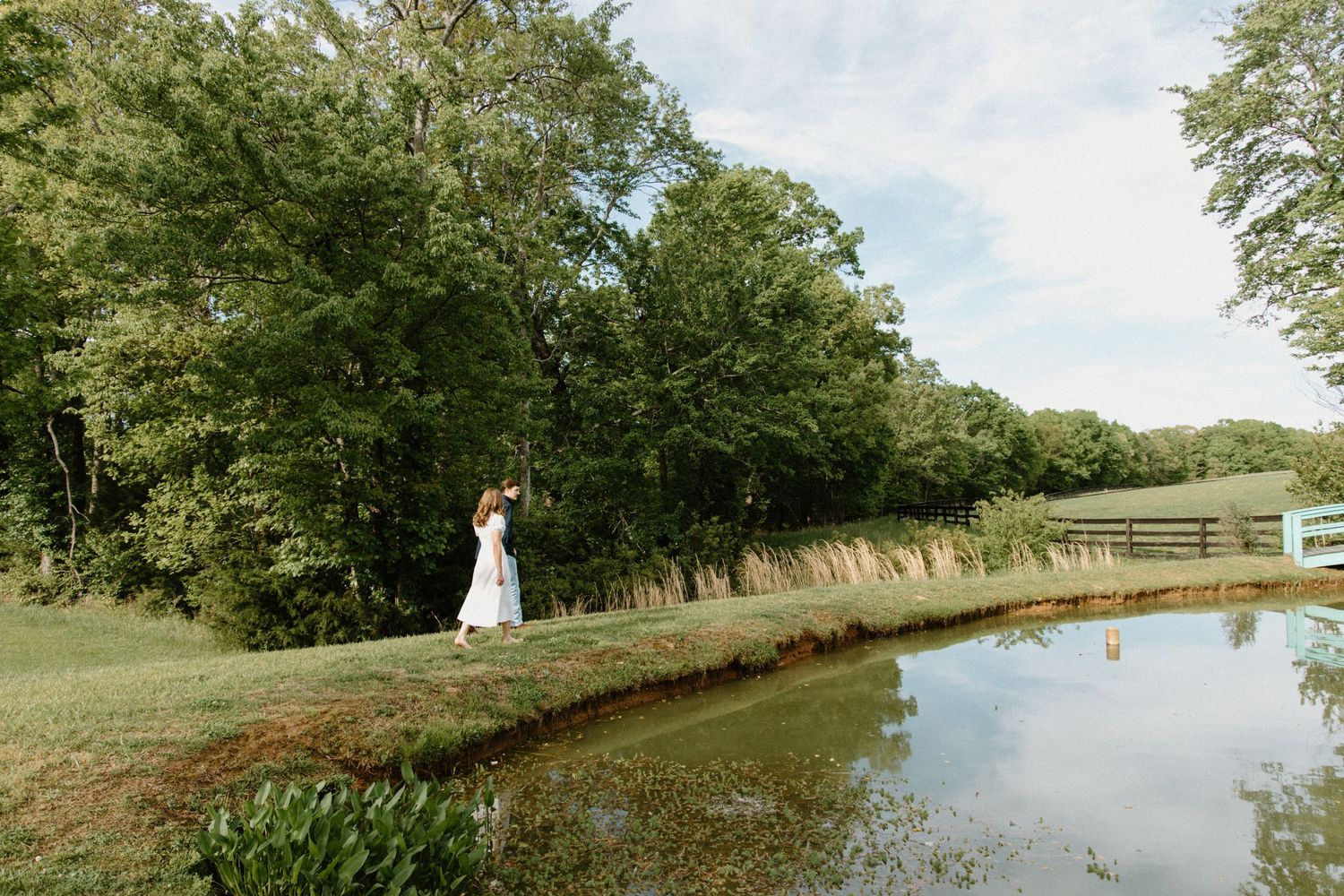 A scenic pond with grassy banks surrounded by lush trees and a pathway along the shoreline on a serene summer day.