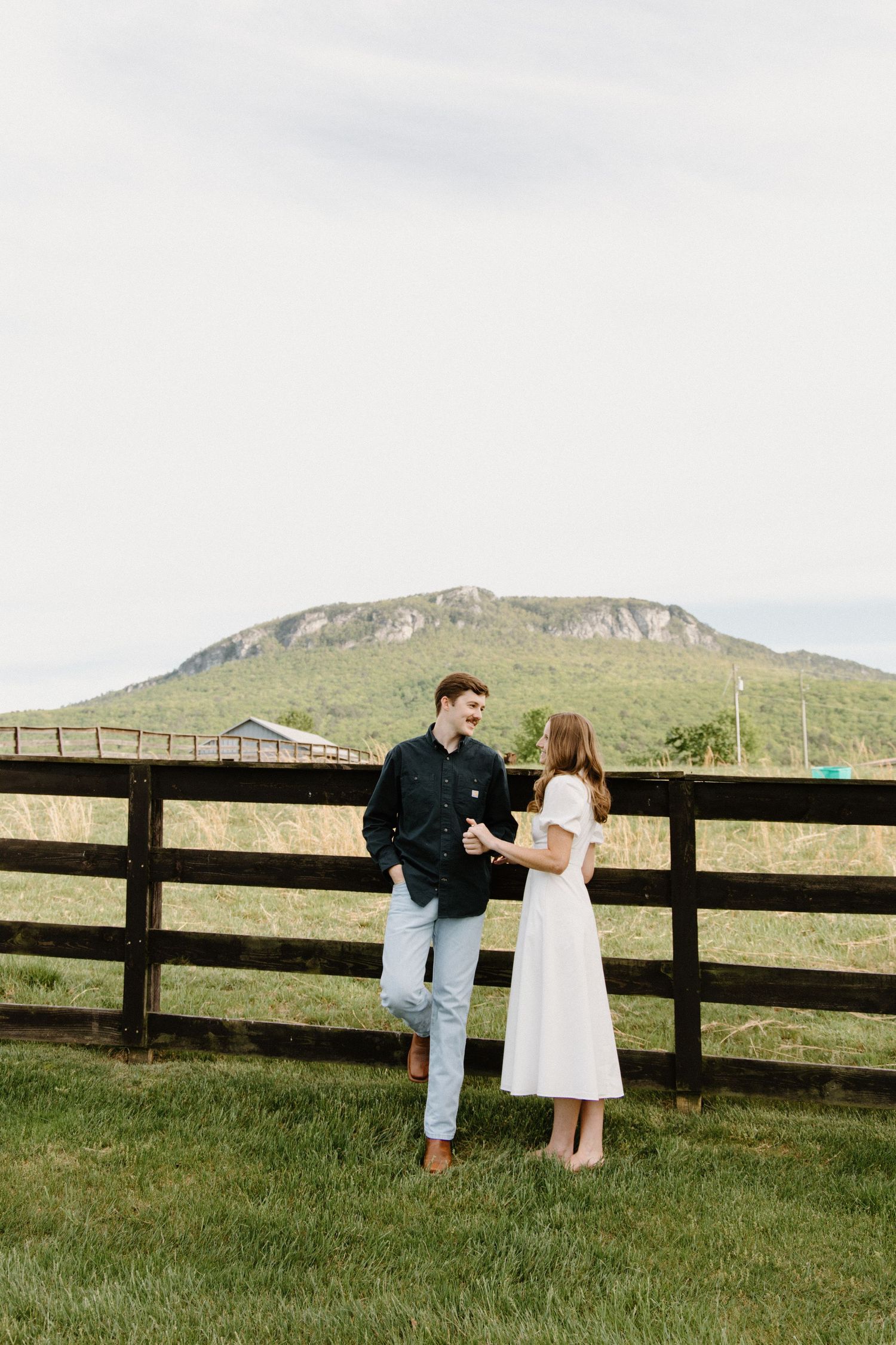 Romantic couple stands by wooden fence overlooking scenic mountain landscape in countryside during sunset.