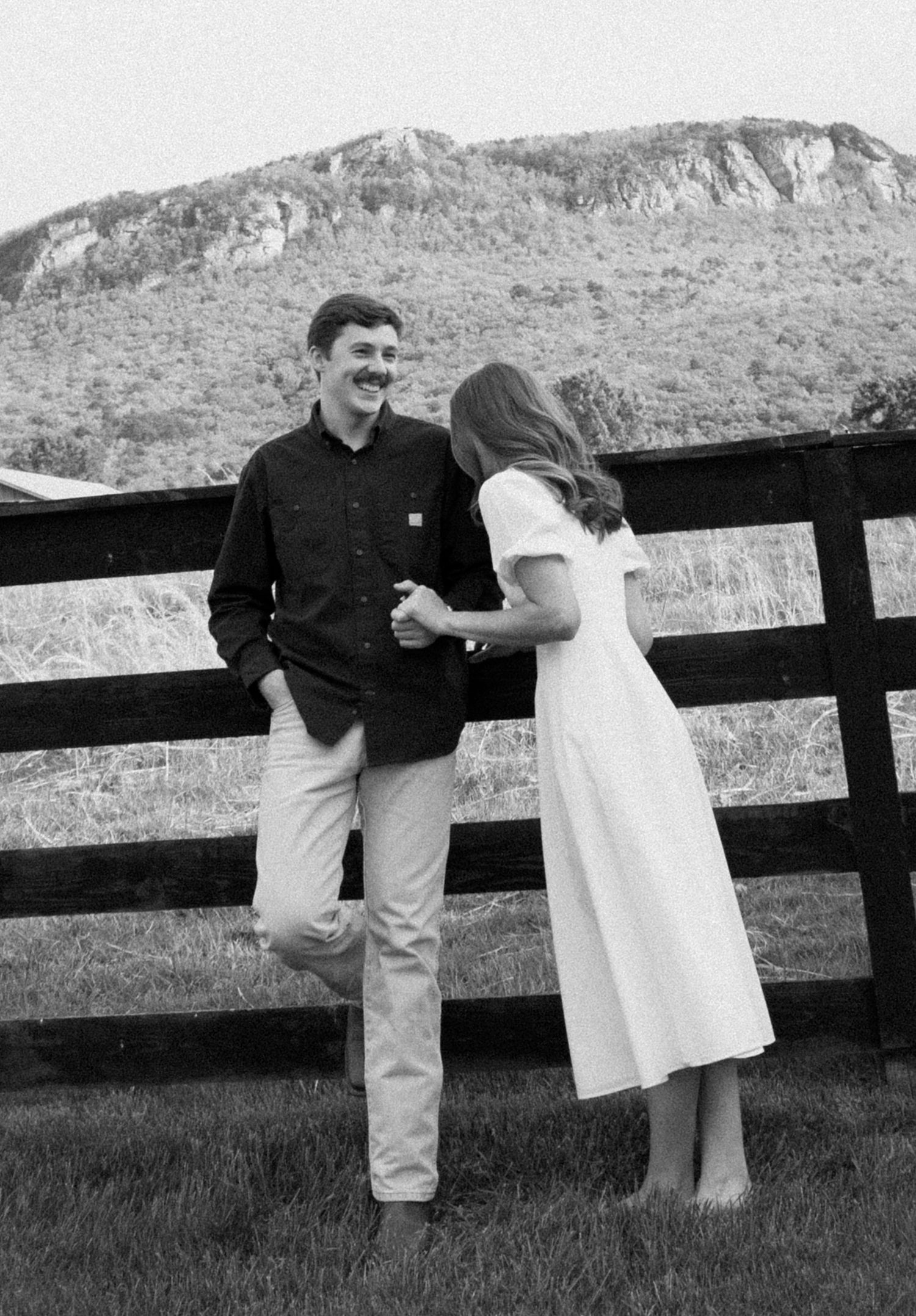 A couple shares a candid black and white moment by a wooden fence with a mountain landscape in the background.
