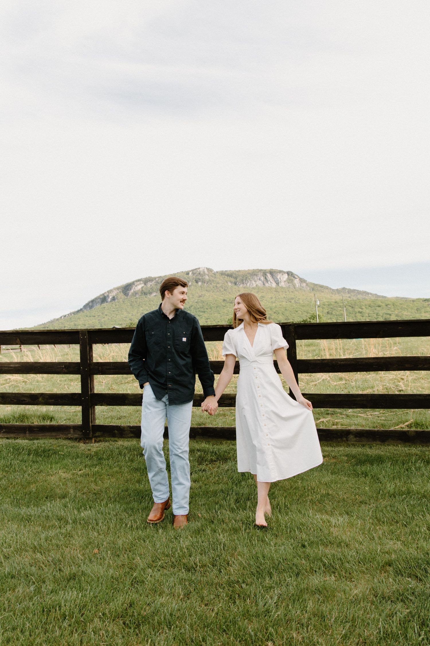 A couple in casual clothing walking together near a rustic wooden fence with rolling hills in the background.