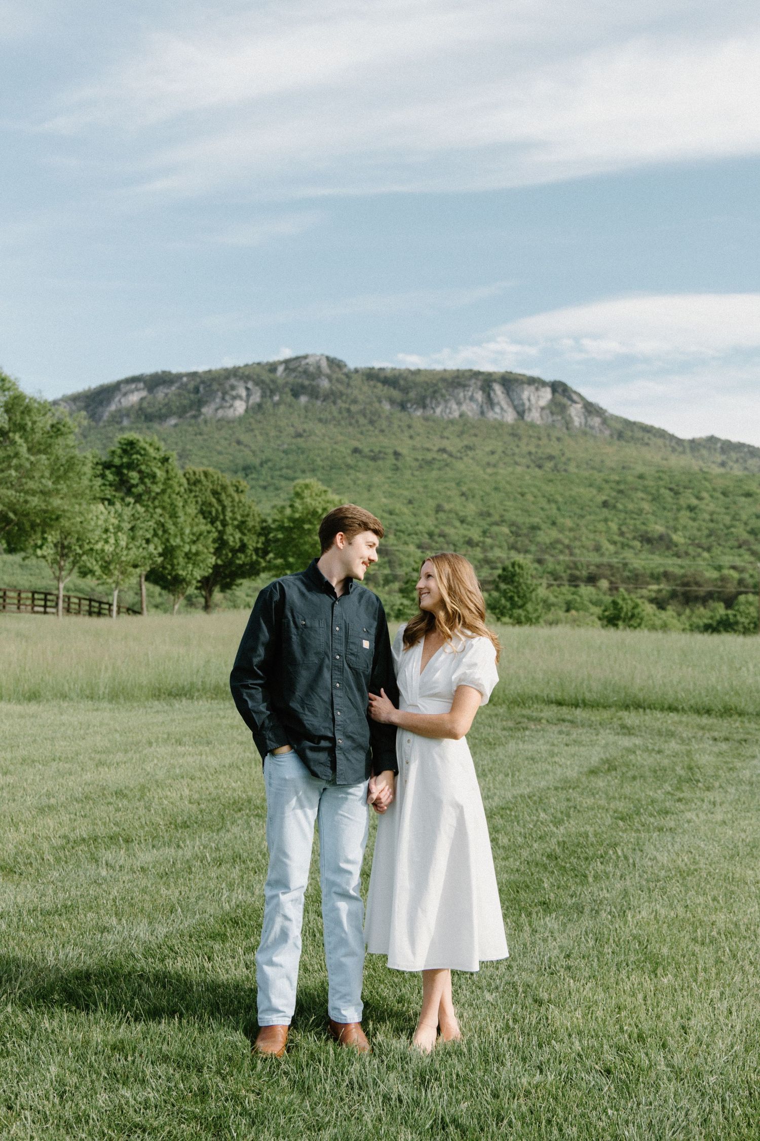 Couple stands together in a scenic field with mountains in background during springtime engagement photoshoot.