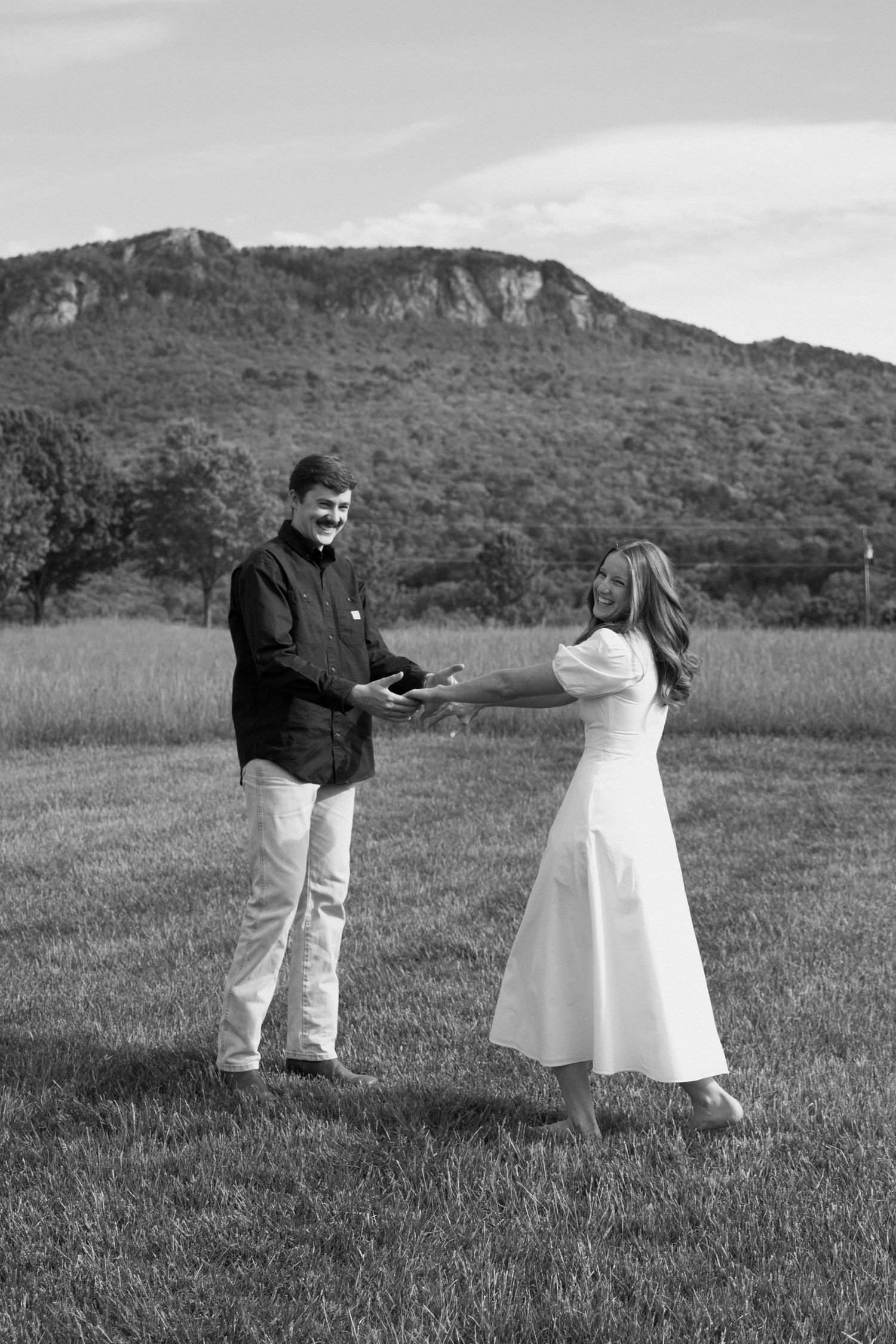 A couple shares a romantic moment in a field with a majestic mountain backdrop in black and white photography.