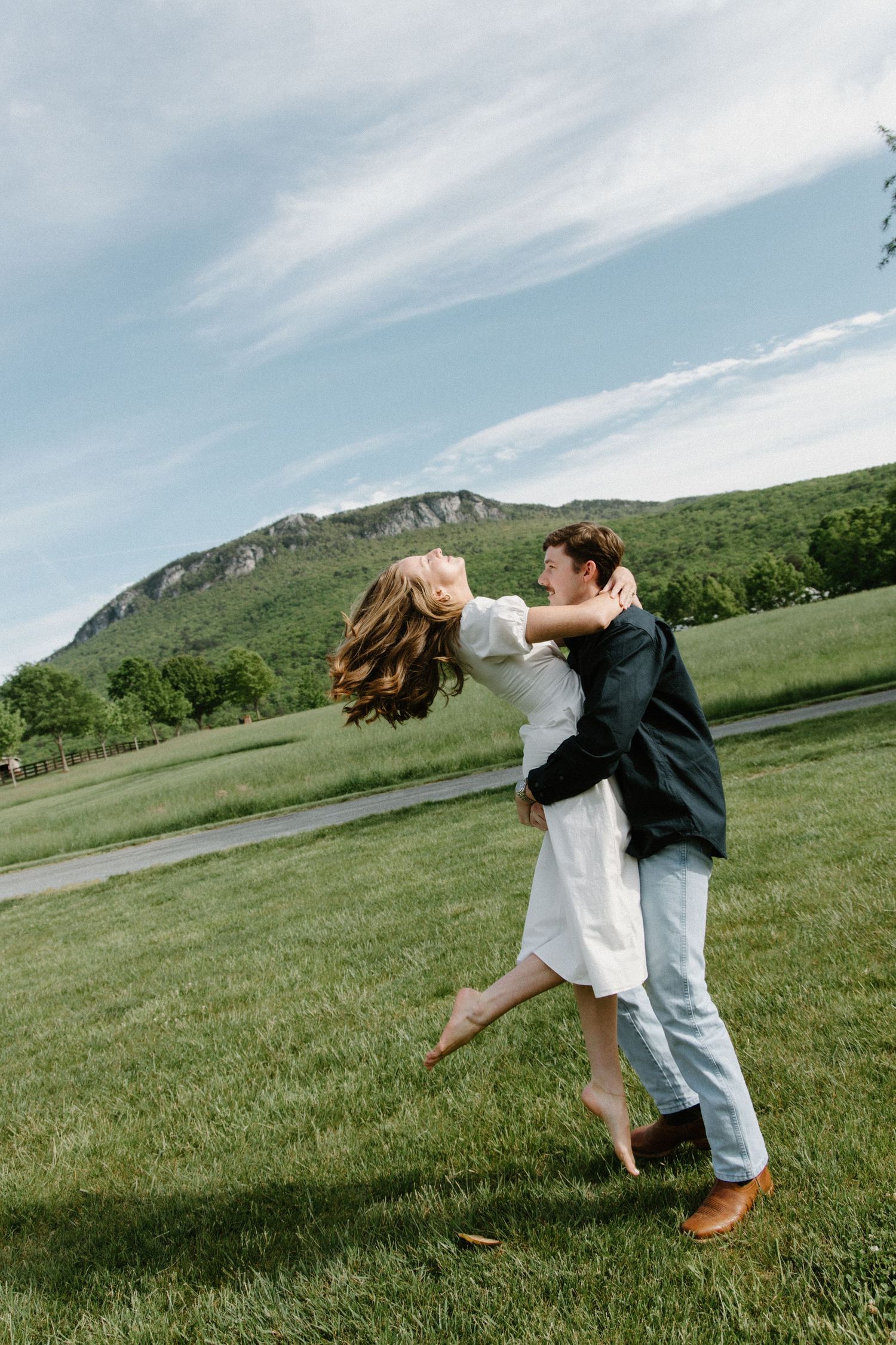 A couple shares a romantic embrace while dancing in a grassy meadow with mountains in the background.