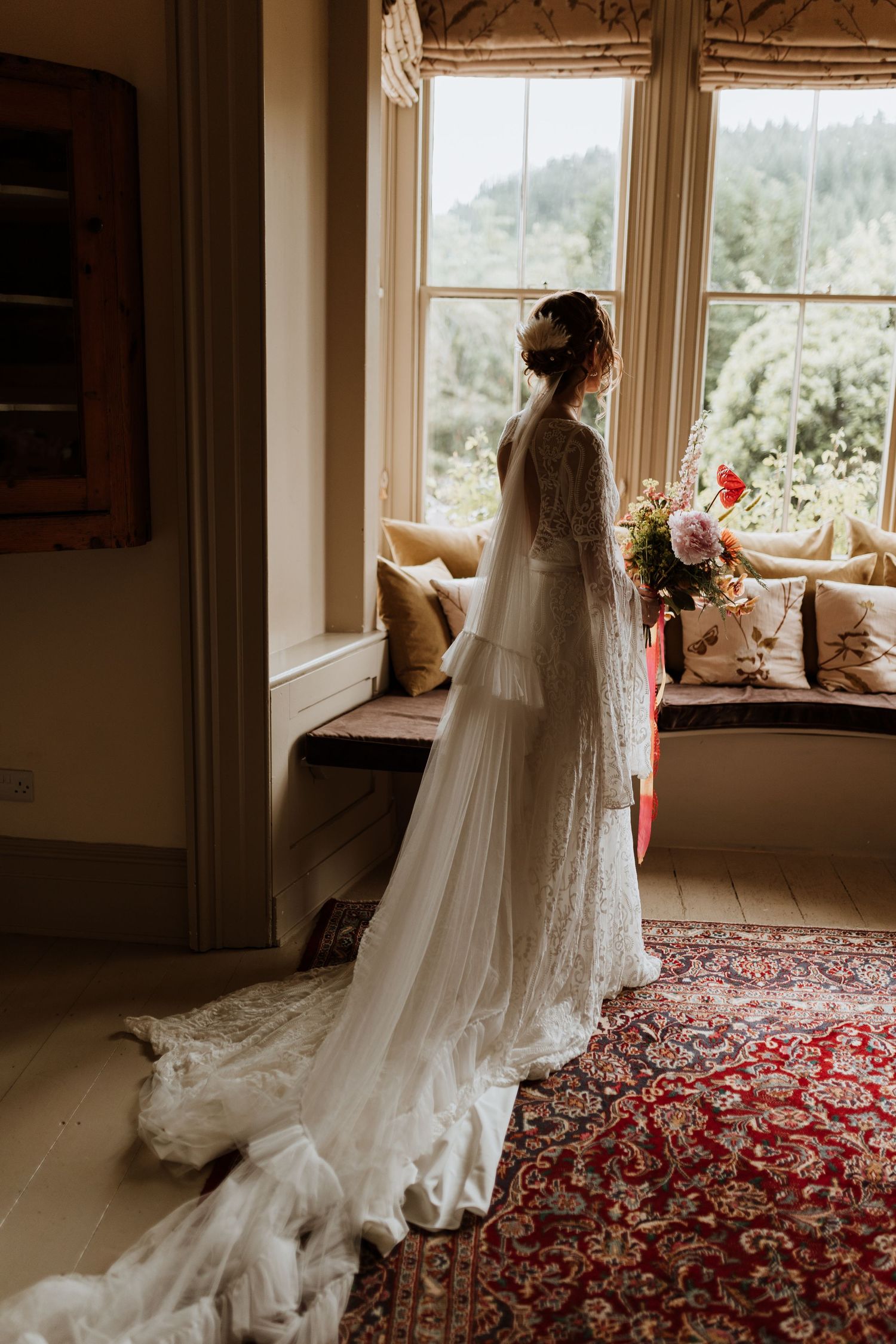 A bride in a flowing white wedding dress with long veil stands by a sunlit window in an elegantly decorated room.
