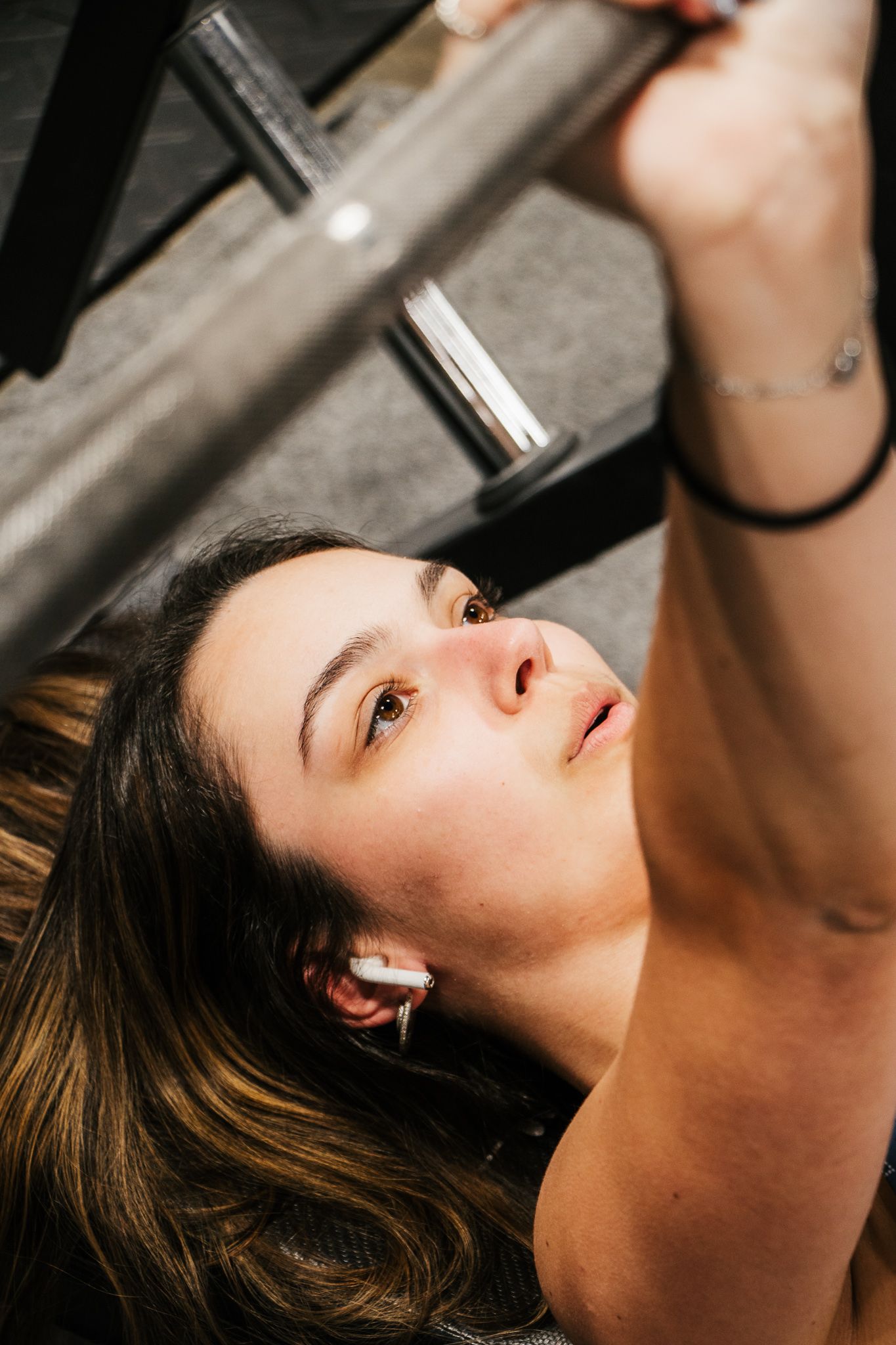 Close-up of a woman lying on a bench, gripping a barbell above her chest and focused during a lift, wearing wireless earbuds.