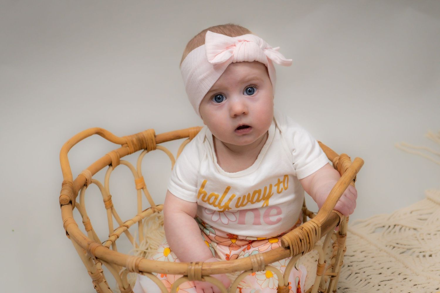 A baby wearing a pink headband and white t-shirt sits in a woven rattan chair against a light background.