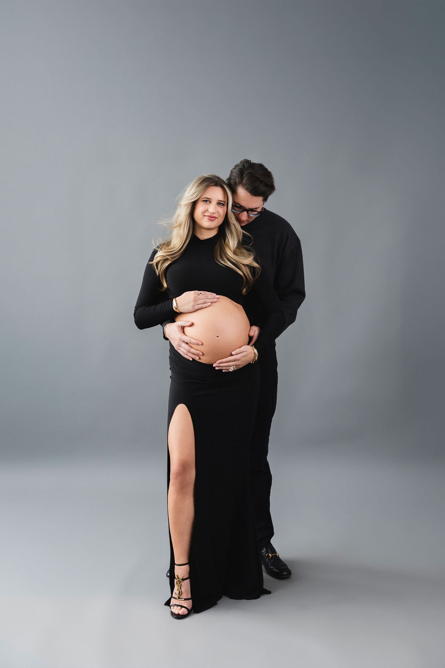 A romantic maternity photoshoot shows couple embracing on a light gray studio backdrop for an elegant maternity portrait.