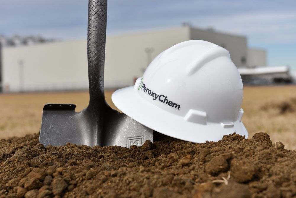 White hard hat with PeroxyChem logo next to shovel on dirt ground against industrial backdrop.