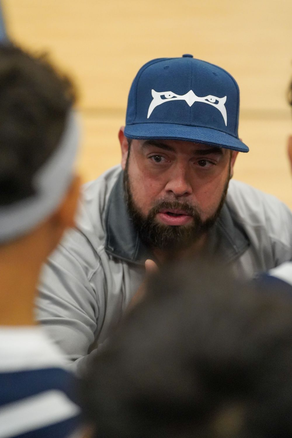 Coach in blue baseball cap with white logo and gray sweatshirt speaking to team during meeting.