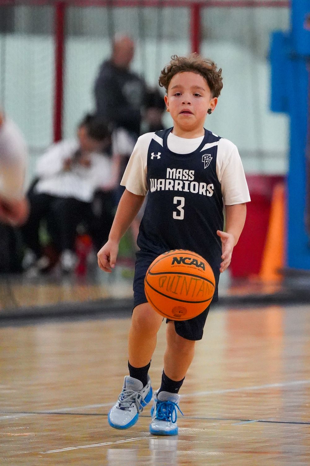 Young basketball player in navy Warriors uniform dribbles on wooden court while practicing ball handling skills.