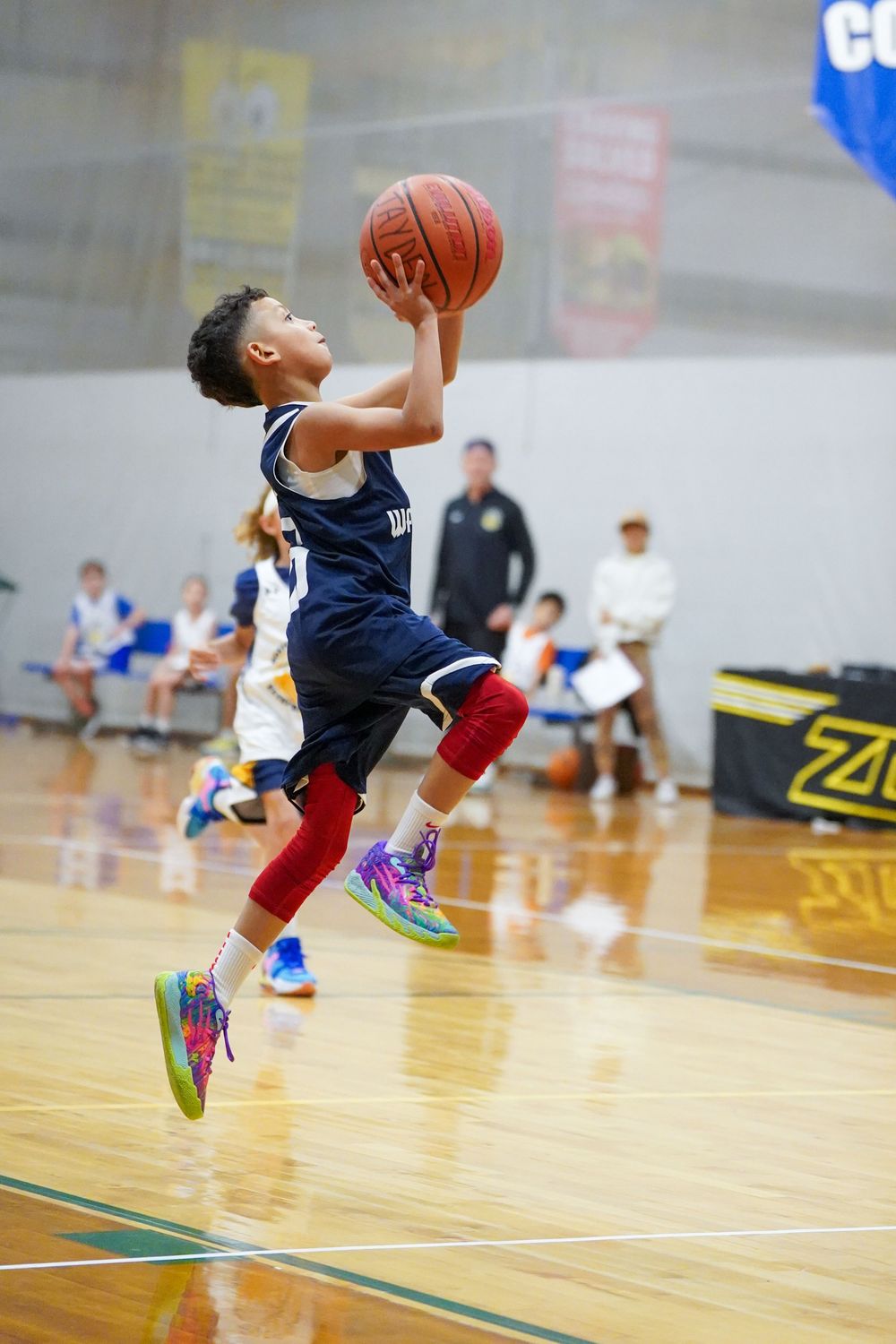 Young basketball player in navy jersey and red socks leaps for layup during indoor youth game on wooden court.