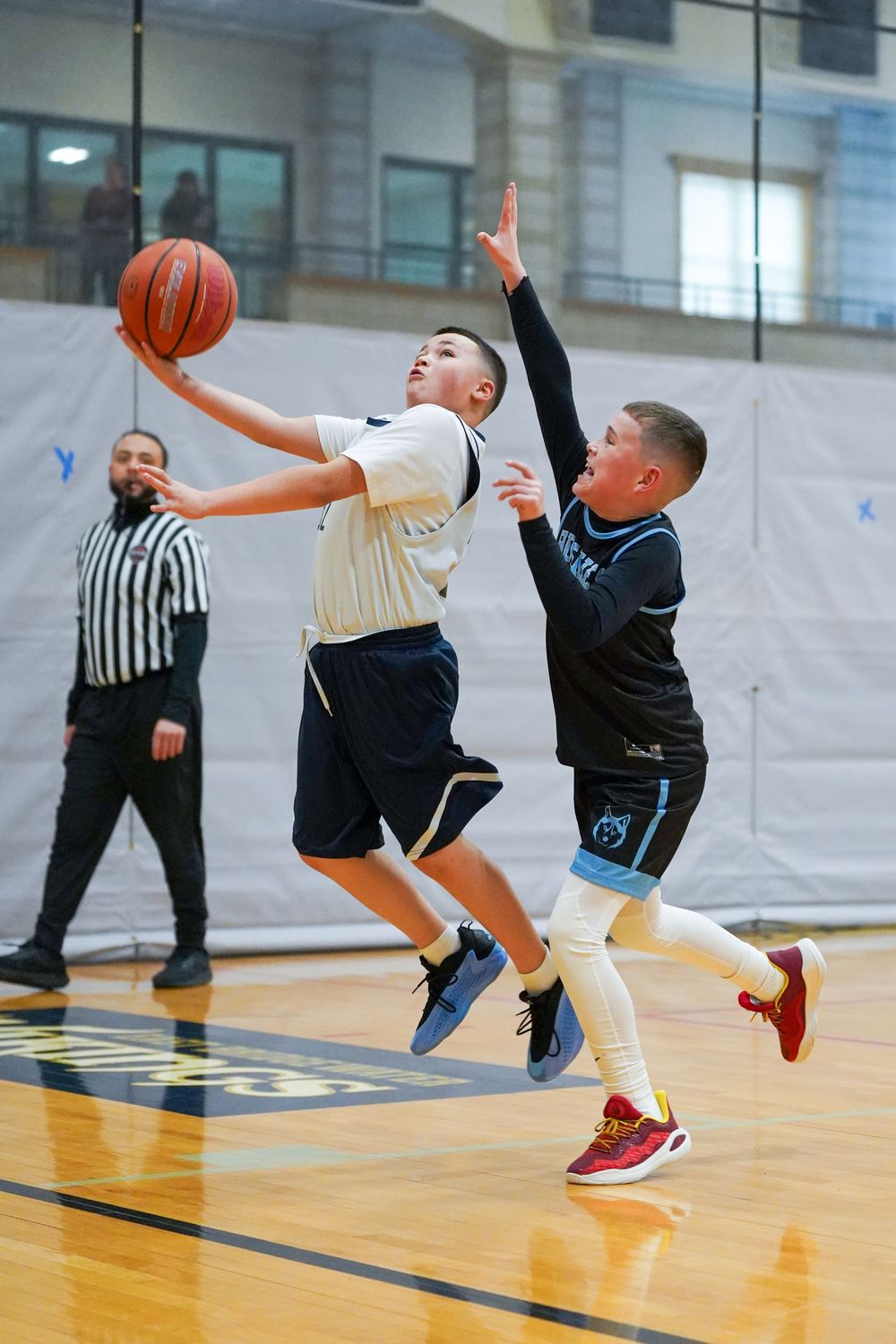 Two youth basketball players compete for a layup during a game while a referee watches from the sideline.