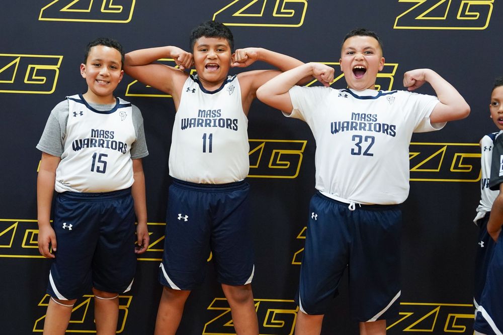 Three basketball players in white jerseys and navy shorts pose playfully with hands behind their ears against a black backdrop.