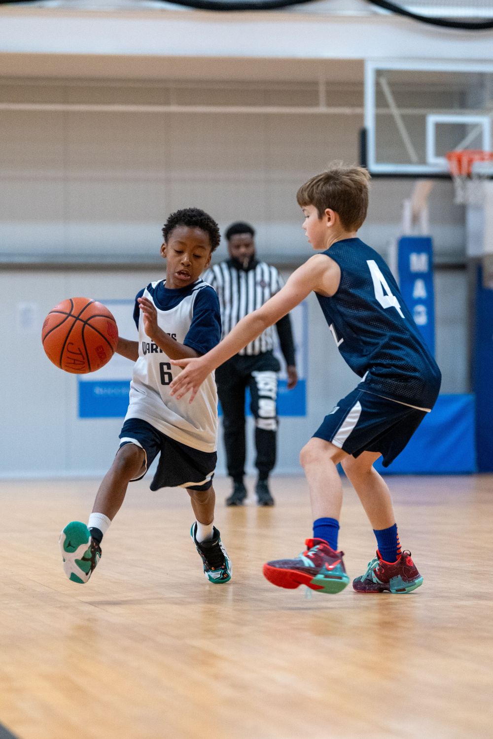 Two young basketball players compete during a youth basketball game on an indoor court with a referee in the background.